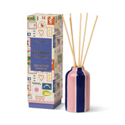 A box labeled “Rosewood Vanilla - A Dopo 4 oz Diffuser” with colorful, whimsical illustrations stands beside a striped ceramic diffuser bottle filled with reed sticks.