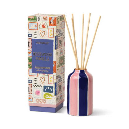 A box labeled “Rosewood Vanilla - A Dopo 4 oz Diffuser” with colorful, whimsical illustrations stands beside a striped ceramic diffuser bottle filled with reed sticks.