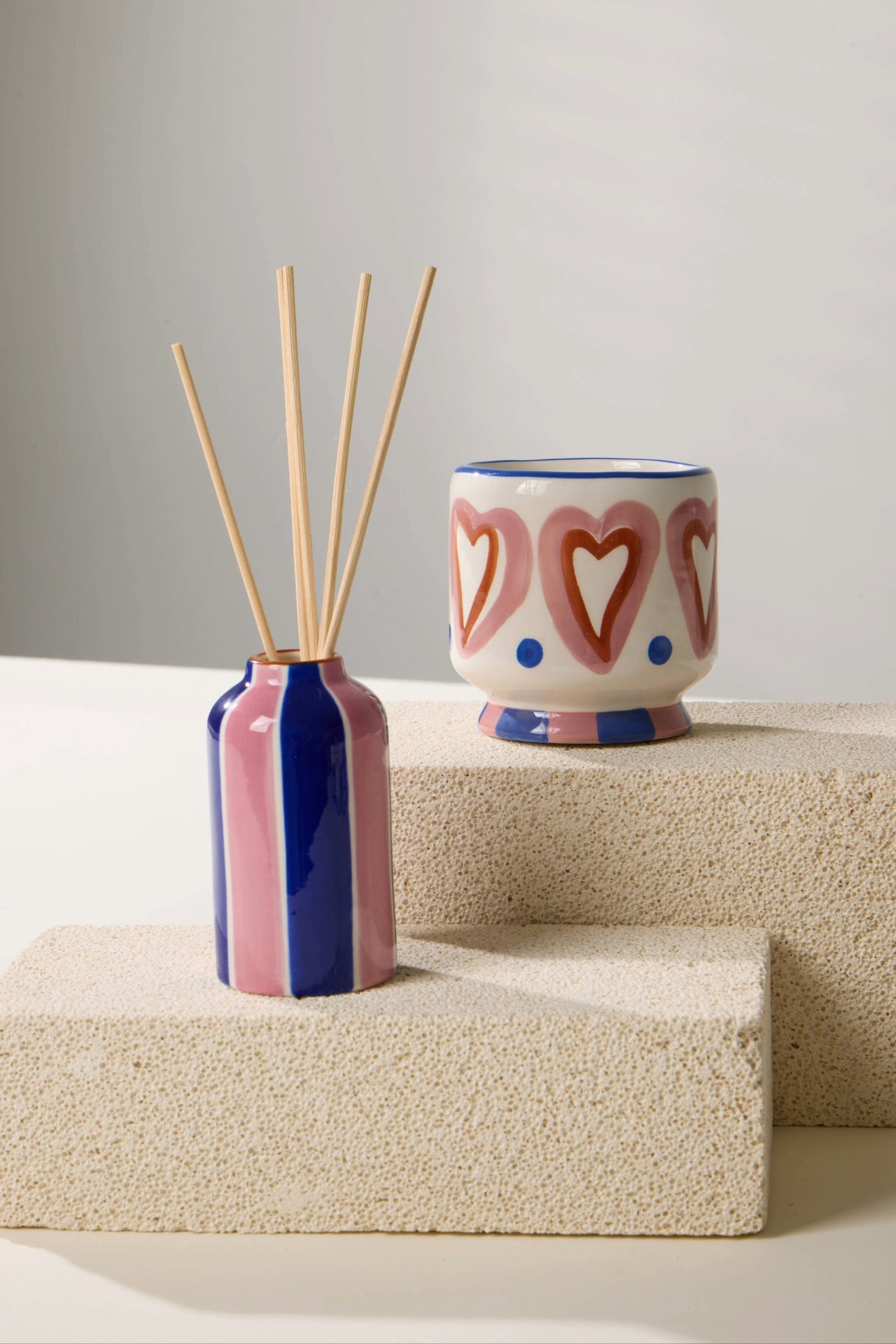 The Rosewood Vanilla - A Dopo 4 oz Diffuser with wooden sticks sits on a textured block beside a ceramic cup with pink hearts and blue dots, both arranged on light-colored surfaces.