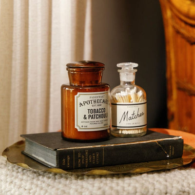 Two vintage apothecary bottles, one labeled Tobacco & Patchouli and the other Matches, sit on a tray atop a book, next to a wicker chair and a soft, textured surface in warm lighting.