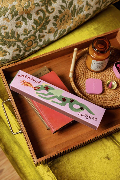 A pink box labeled Dragon - Extra Long Boxed Matches with a green snake sits on a tray alongside books, a candle, and more atop a yellow-green upholstered surface.