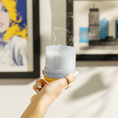 A hand with silver nail polish holds a blue candle with smoke rising from its extinguished wick, in front of framed art on a wall.