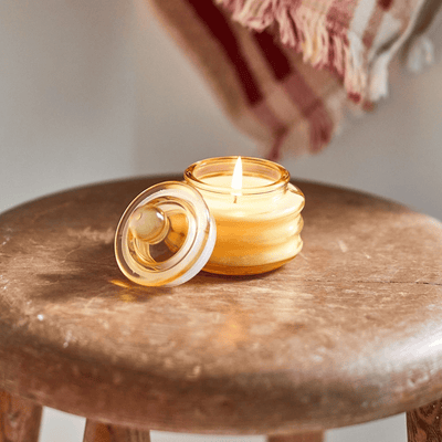 A small, yellow glass candle jar with its lid off sits on a rustic wooden stool. The candle is lit, and a striped fabric is visible in the blurred background.