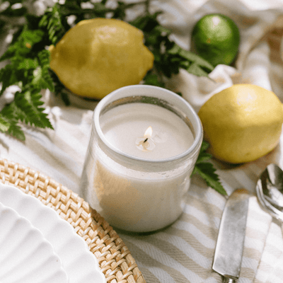 A lit white candle in a glass jar sits on a striped cloth near lemons, a lime, greenery, a woven placemat, a white plate, a knife, and a spoon, creating a fresh, summery table setting.