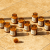 Twelve amber glass jars with white labels are arranged on a tan tiled surface; one jar lies on its side with the lid off. Sunlight casts shadows across the scene.