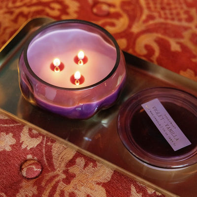 A purple glass candle with four lit wicks sits on a gold tray next to its lid. The tray rests on a red and gold patterned fabric.
