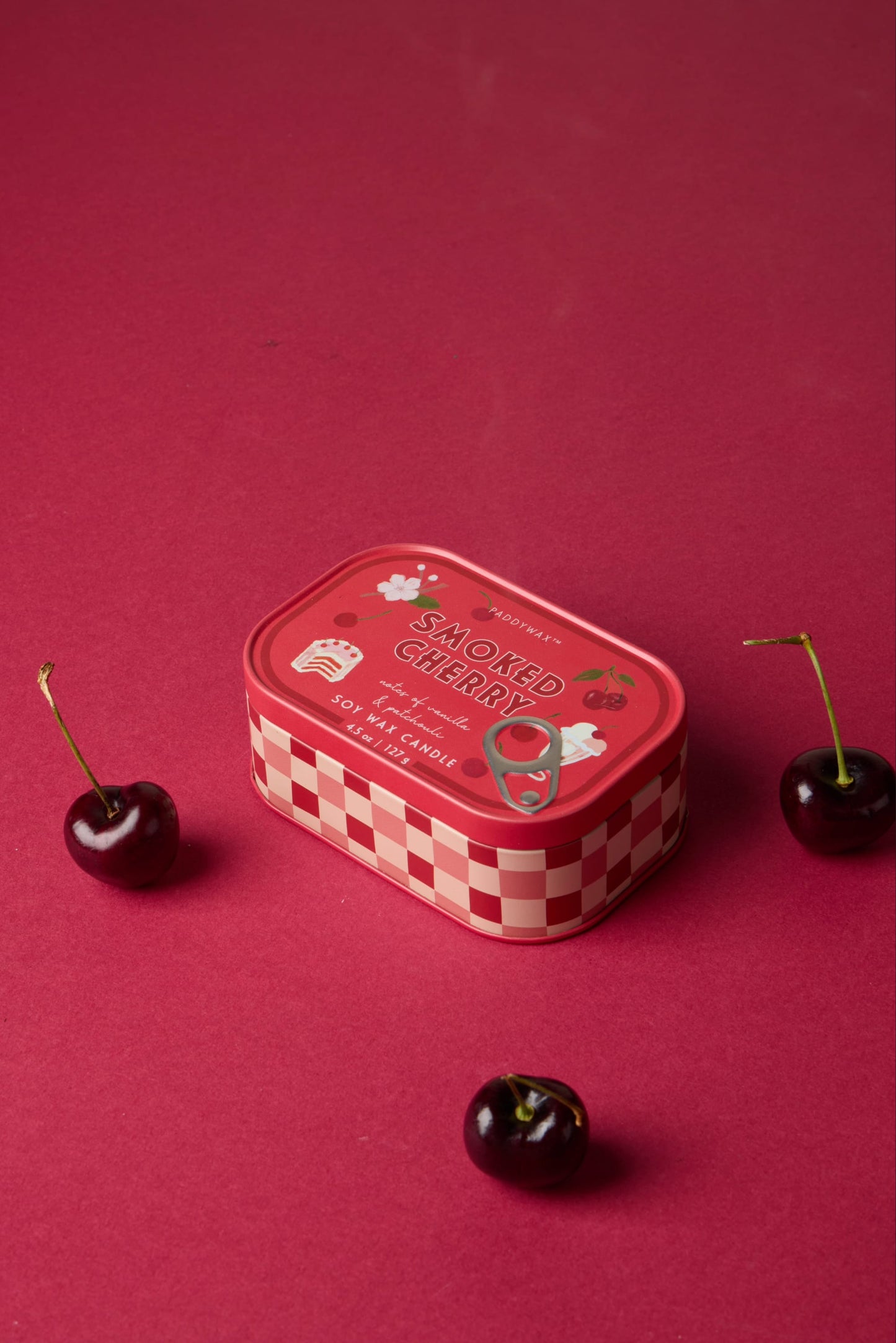 A Smoked Cherry - Bistro Tin 4.5 oz Candle, with a red checkered pattern and cherry illustrations, rests on a red surface next to three fresh cherries.