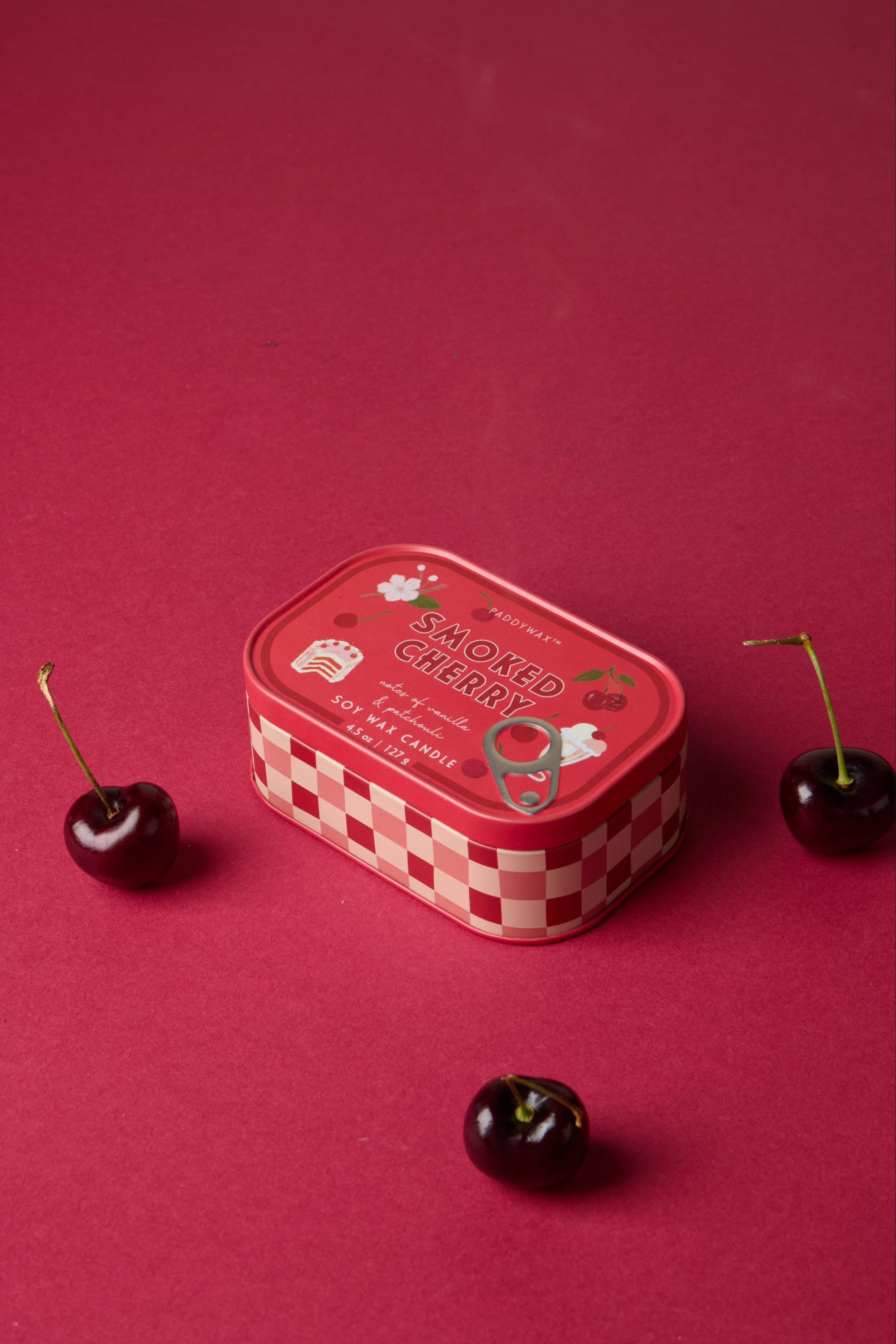 A Smoked Cherry - Bistro Tin 4.5 oz Candle, with a red checkered pattern and cherry illustrations, rests on a red surface next to three fresh cherries.