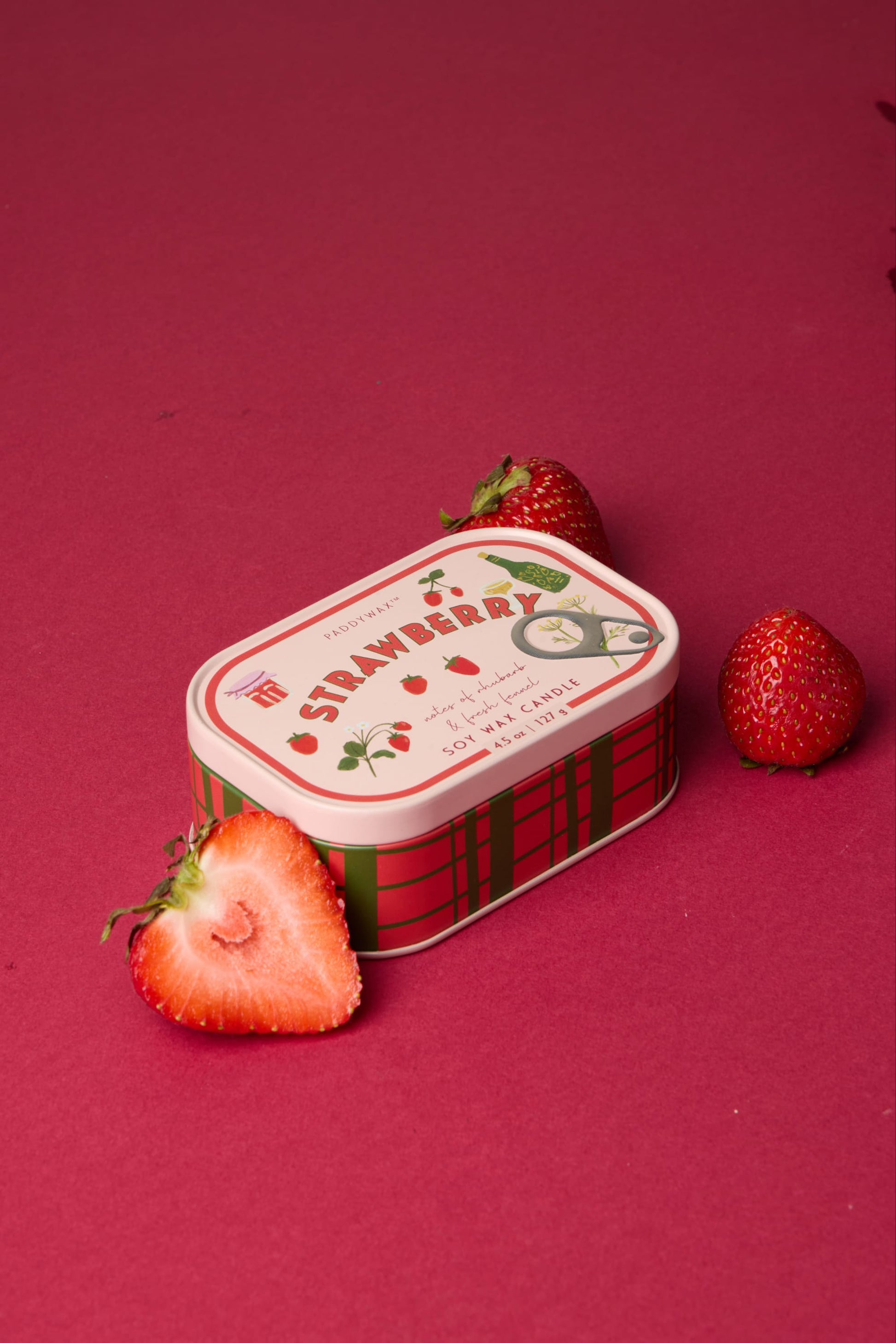 A Strawberry - Bistro Tin 4.5 oz Candle, with "Strawberry" on its lid and strawberry illustrations, sits on a red surface surrounded by three strawberries, one sliced in half.