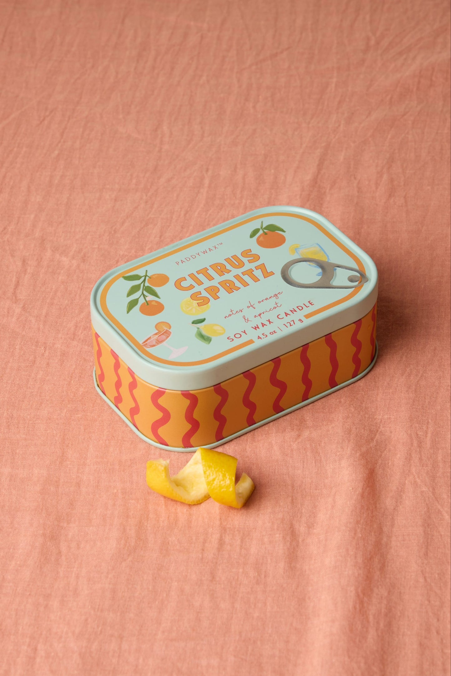 A colorful Citrus Spritz - Bistro Tin 4.5 oz Candle, decorated with fruit illustrations, rests on peach fabric. A small curl of lemon peel is placed in front of the tin.