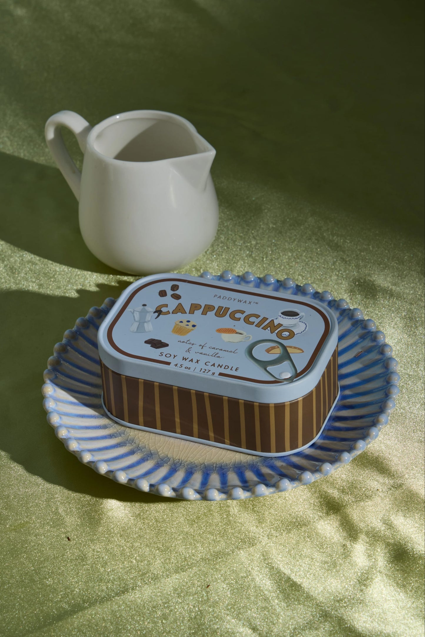A small white pitcher and a Cappuccino - Bistro 4.5 oz Tin Candle rest on a blue-rimmed plate atop a green textured surface, bathed in natural sunlight that casts gentle shadows.