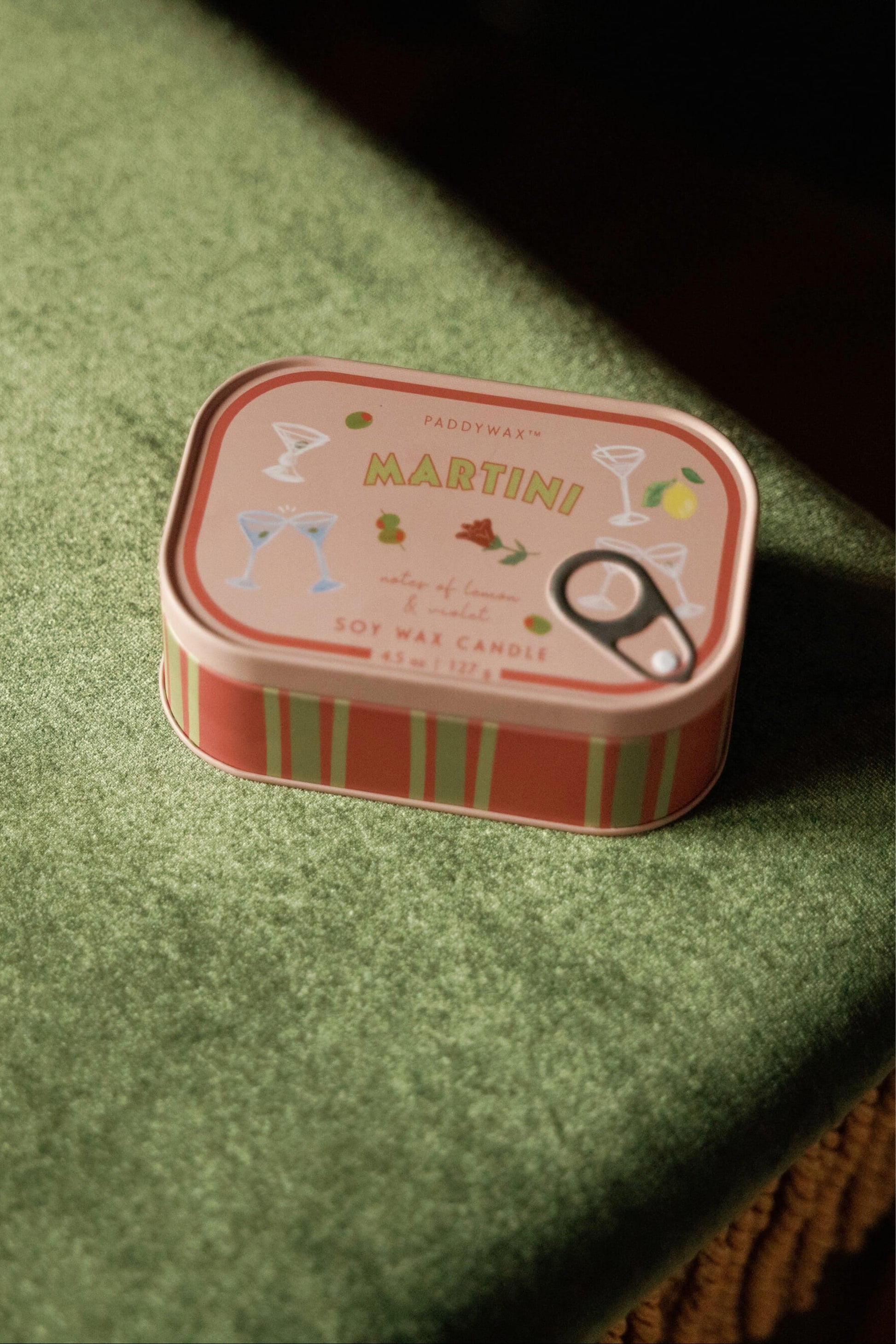 A Martini - Bistro 4.5 oz Tin Candle, featuring cocktail glass illustrations on its pink tin, rests on a green velvet surface.