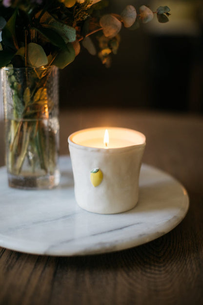 The Meyer Lemon - Bistro 8 oz Candle glows with a citrus scent on a round marble tray beside a glass vase of flowers—an elegant gift candle for any wooden table.