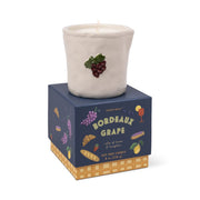 The Bordeaux Grape - Bistro 8 oz Candle by Paddywax is a white aromatic soy candle with grape cluster decoration, resting on a blue box featuring images of grapes, bread, and cheese, and offers notes of blossom and honeysuckle.