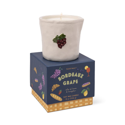 The Bordeaux Grape - Bistro 8 oz Candle by Paddywax is a white aromatic soy candle with grape cluster decoration, resting on a blue box featuring images of grapes, bread, and cheese, and offers notes of blossom and honeysuckle.