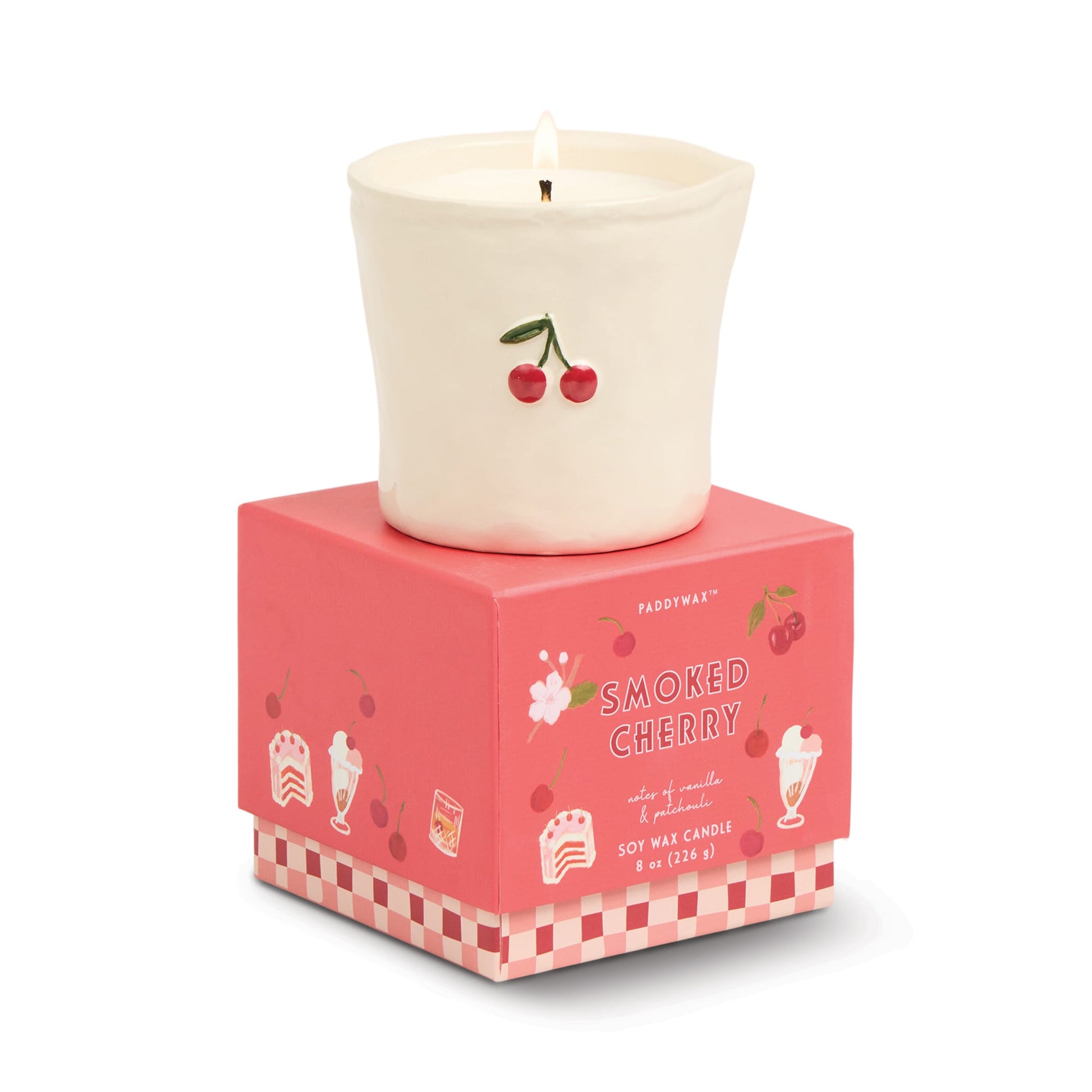 A lit white ceramic candle with a cherry design sits on a pink box labeled "Smoked Cherry - Bistro 8 oz Candle," featuring cherries and dessert illustrations, making this soy wax candle both stylish and inviting.