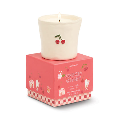 A lit white ceramic candle with a cherry design sits on a pink box labeled "Smoked Cherry - Bistro 8 oz Candle," featuring cherries and dessert illustrations, making this soy wax candle both stylish and inviting.
