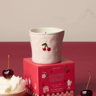 A white ceramic candle holder with a cherry design sits atop the Smoked Cherry - Bistro 8 oz Candle box. Nearby are fresh cherries and whipped cream on a red and cream backdrop. The holder is food safe for reuse.