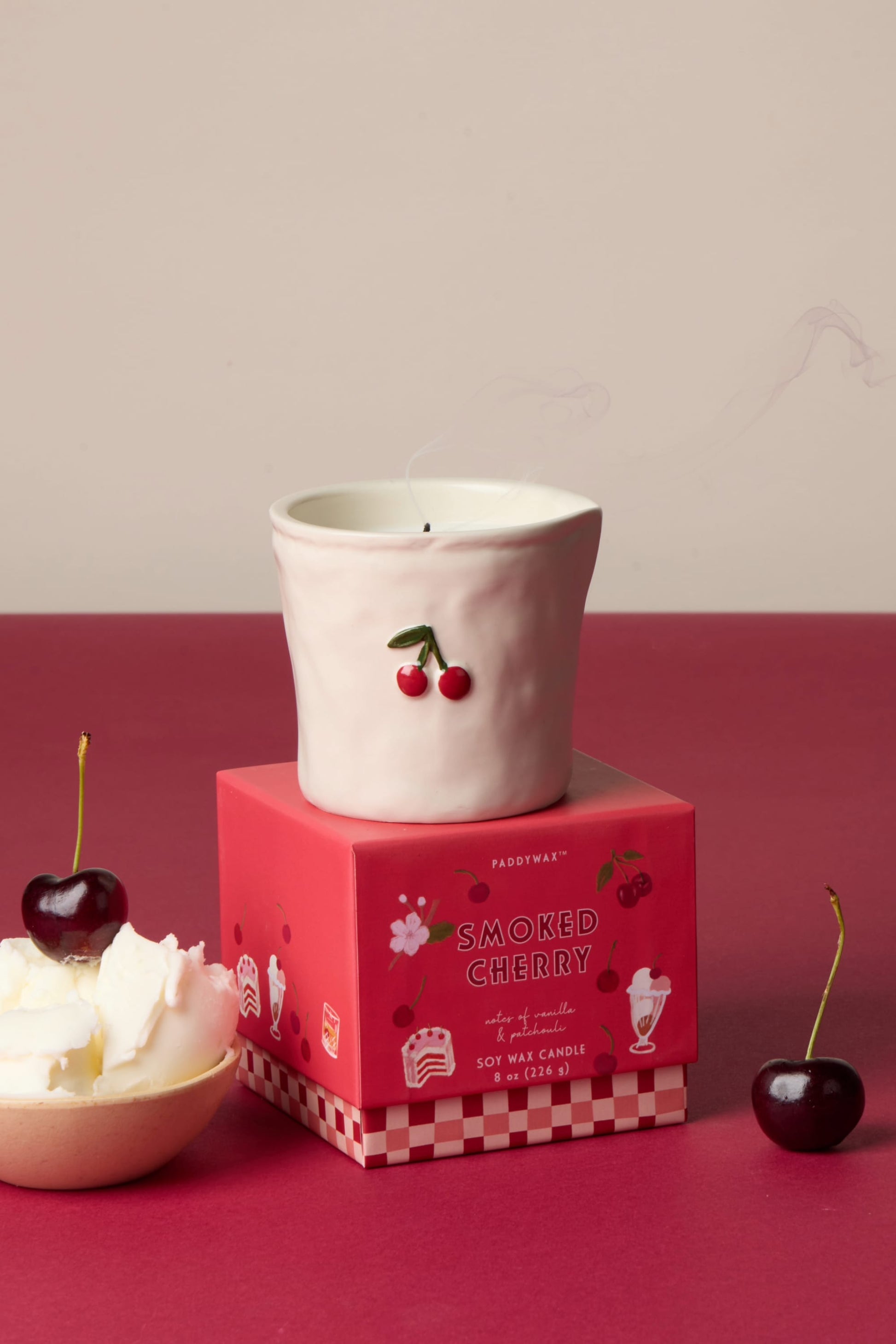 A white ceramic candle holder with a cherry design sits atop the Smoked Cherry - Bistro 8 oz Candle box. Nearby are fresh cherries and whipped cream on a red and cream backdrop. The holder is food safe for reuse.