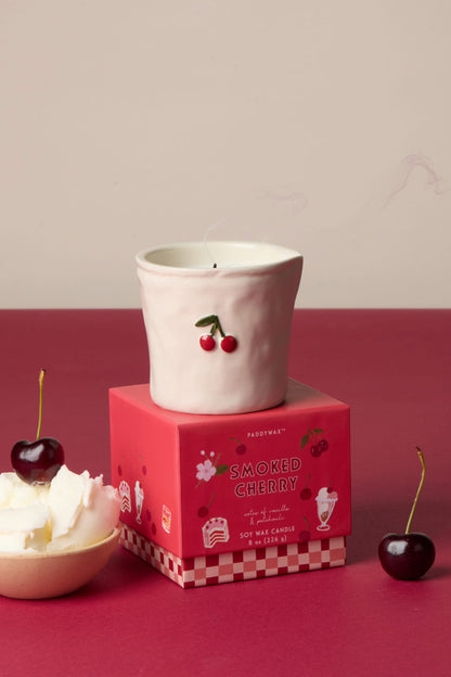 A white ceramic candle holder with a cherry design sits atop the Smoked Cherry - Bistro 8 oz Candle box. Nearby are fresh cherries and whipped cream on a red and cream backdrop. The holder is food safe for reuse.
