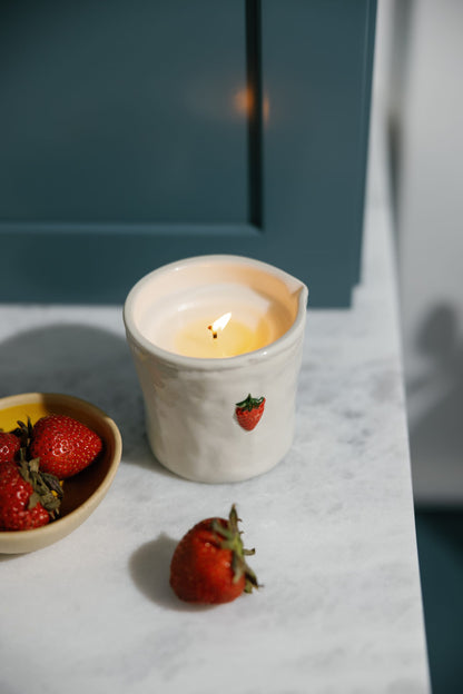 The Strawberry - Bistro 8 oz Candle, in a white ceramic holder with a strawberry design, is displayed on marble next to a bowl of strawberries and a single berry beside it.