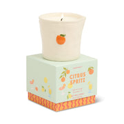 The Citrus Spritz - Bistro 8 oz Candle, a white ceramic candle topped with an orange fruit accent, sits on a pastel green box adorned with citrus art, vibrant drinks, and orange-pink wavy patterns along the bottom.