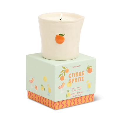 The Citrus Spritz - Bistro 8 oz Candle, a white ceramic candle topped with an orange fruit accent, sits on a pastel green box adorned with citrus art, vibrant drinks, and orange-pink wavy patterns along the bottom.