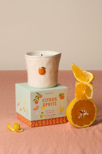 The Citrus Spritz - Bistro 8 oz Candle, a white ceramic candle with an orange fruit design, rests on a pastel box. Nearby are an orange wedge, a lemon wedge, and a spiral lemon peel on a pink surface.