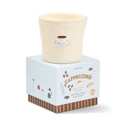 A cream-colored Cappuccino - Bistro 8 oz Candle, infused with a rich coffee aroma, is presented in a charming ceramic bistro cup and sits atop a box adorned with illustrations of coffee and pastries. The reusable ceramic vessel makes it both an eco-friendly choice and a delightful decor piece.