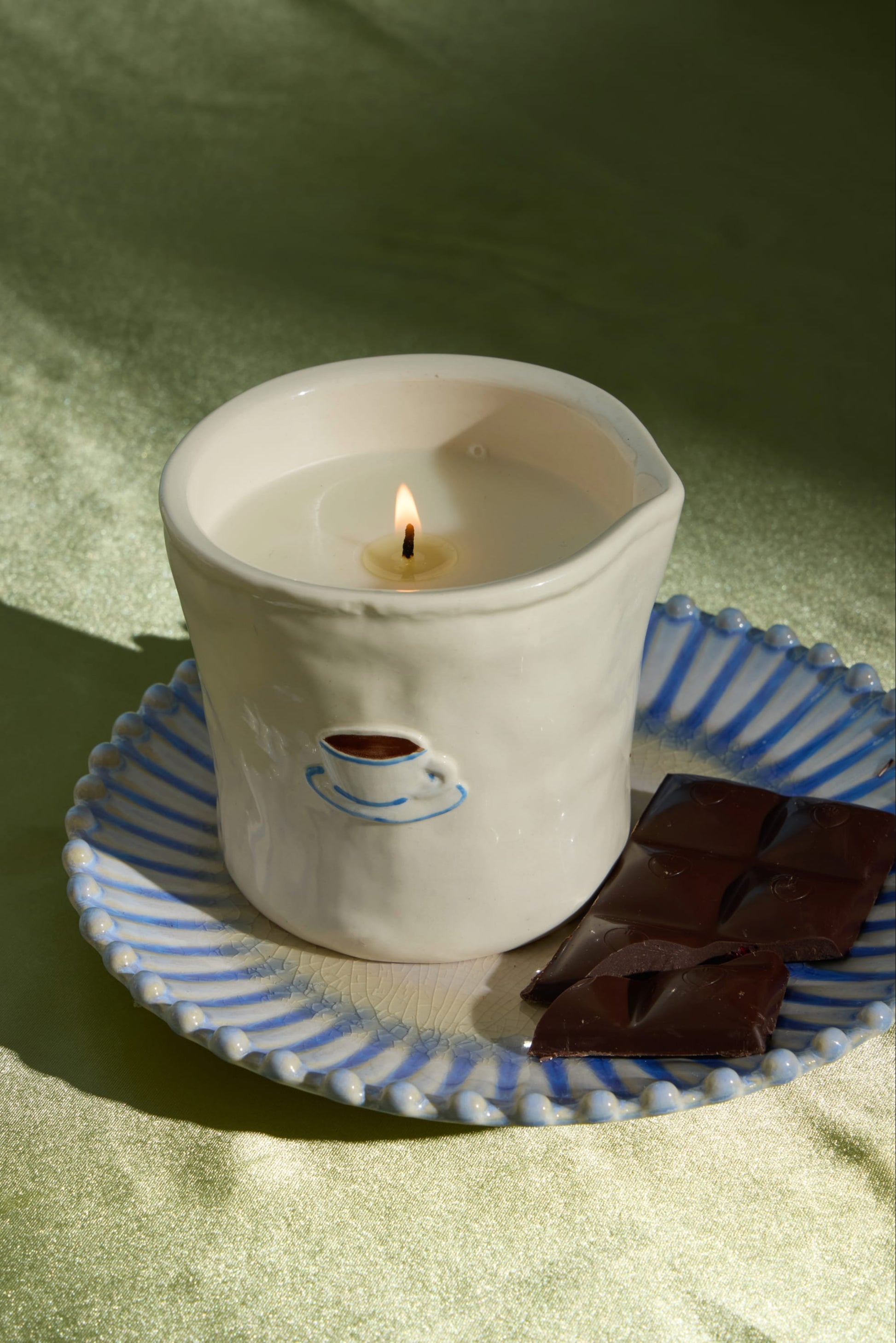 A lit Cappuccino - Bistro 8 oz Candle in a ceramic cup with a coffee cup design sits on a blue-striped plate next to two pieces of dark chocolate on a green surface.