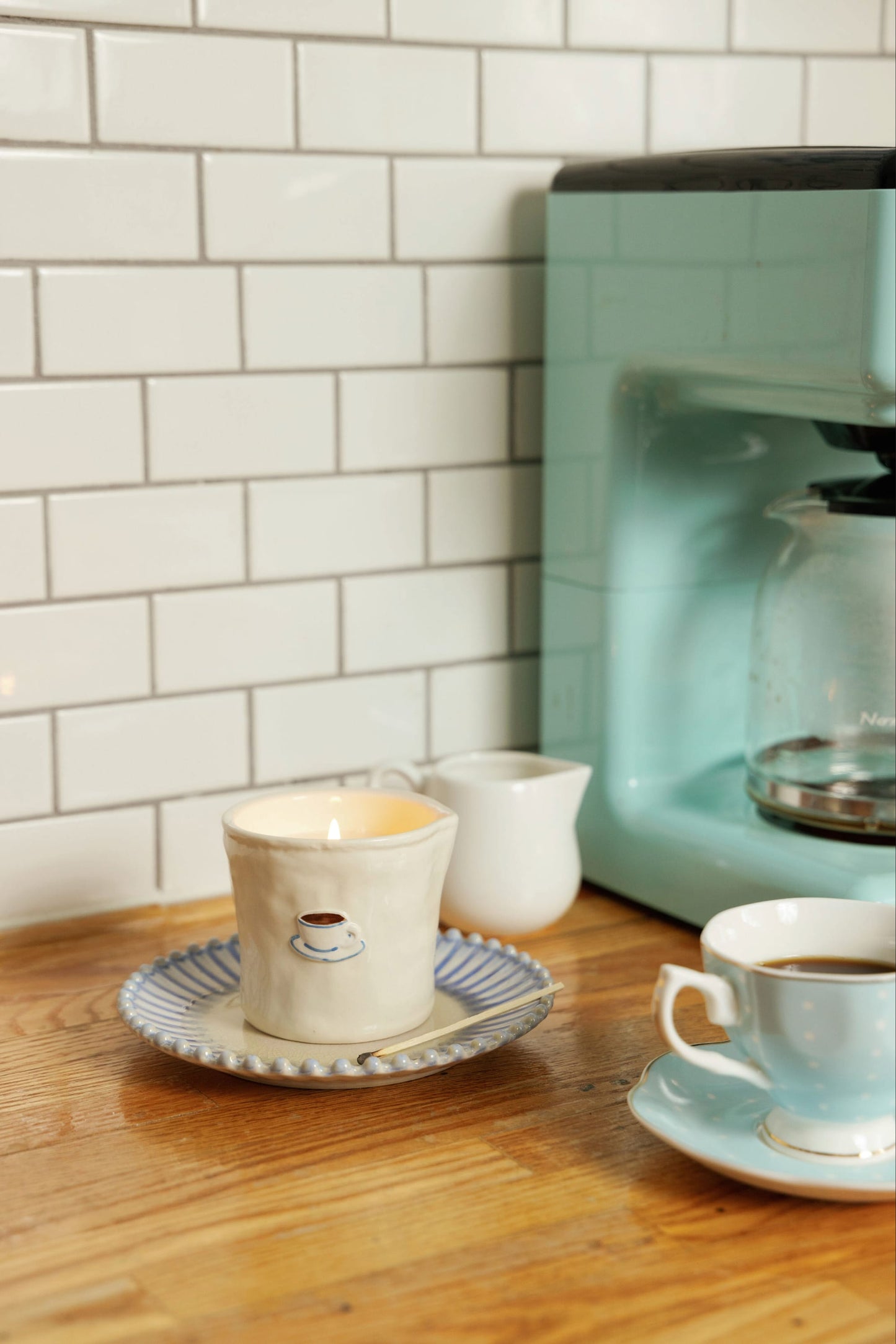A lit Cappuccino - Bistro 8 oz Candle in a ceramic mug sits on a saucer next to a cup of coffee, a small creamer, and a teal coffee maker on a wooden counter with white subway tile backsplash.