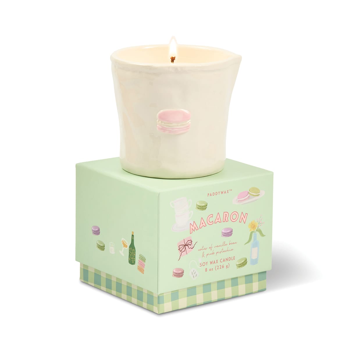 A lit Macaron - Bistro 8 oz Candle, featuring a white ceramic vessel with a pink macaron illustration, sits atop a pastel green macaron-themed box decorated with colorful macaron drawings and text—creating the perfect macaron-inspired candle housed in a charming, food-safe ceramic container.