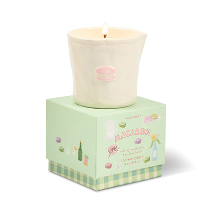 A lit Macaron - Bistro 8 oz Candle, featuring a white ceramic vessel with a pink macaron illustration, sits atop a pastel green macaron-themed box decorated with colorful macaron drawings and text—creating the perfect macaron-inspired candle housed in a charming, food-safe ceramic container.
