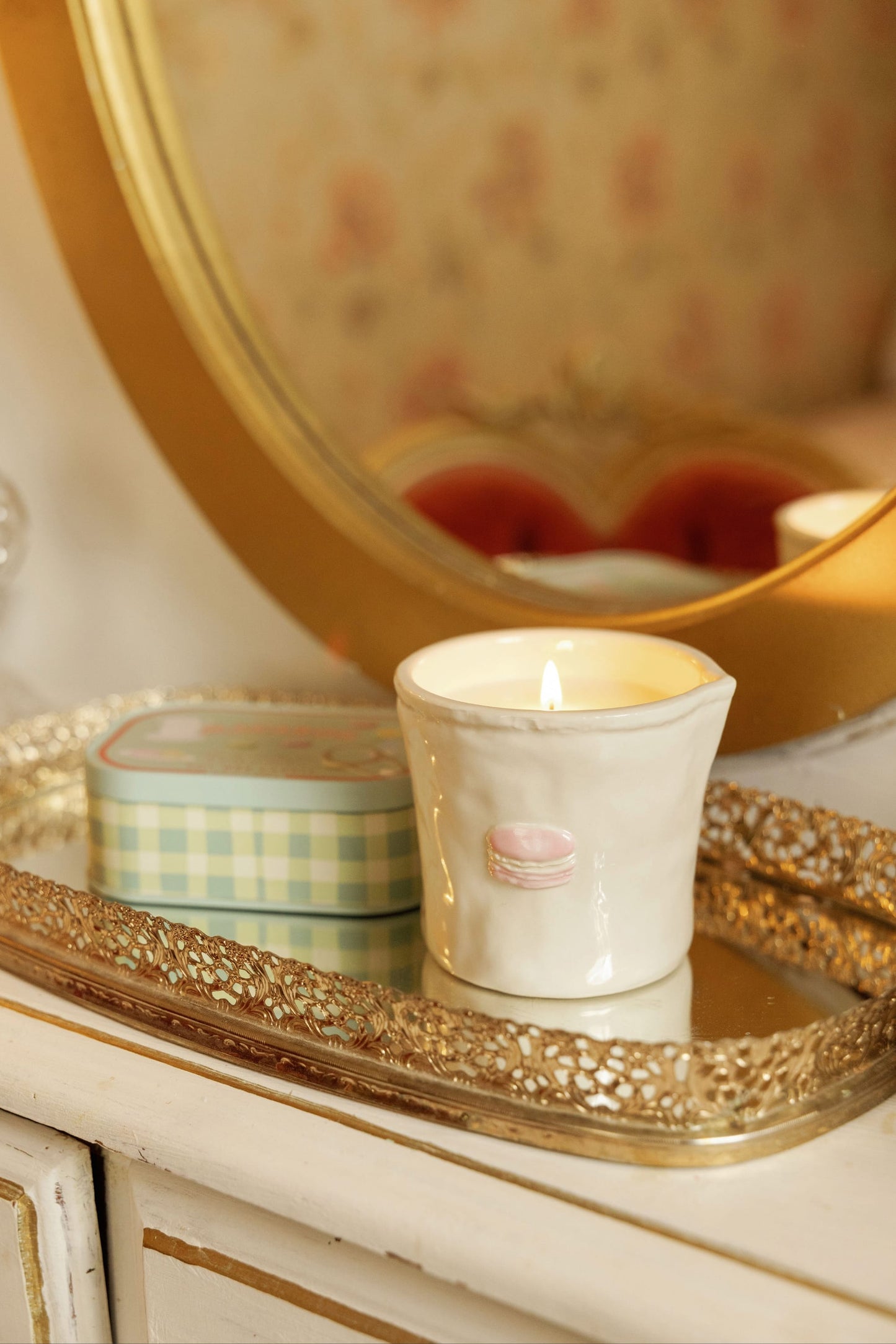 A lit Macaron - Bistro 8 oz Candle sits in a ceramic holder beside a small tin on a decorative gold tray atop a white vanity, with a mirror and soft floral wallpaper in the background.