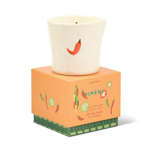 A white ceramic candle holder featuring a red chili pepper motif rests atop an orange box adorned with illustrations of peppers and limes. The bistro-inspired design adds a delightful touch, while the packaging reads “Piment - Bistro 8 oz Candle, soy wax (226 g),” hinting at the distinctive Piment fragrance inside.