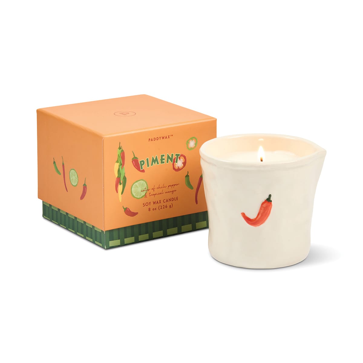 A lit Piment - Bistro 8 oz Candle in a ceramic holder featuring a red chili pepper design sits beside its orange box labeled “Piment,” decorated with vegetable illustrations and green trim.