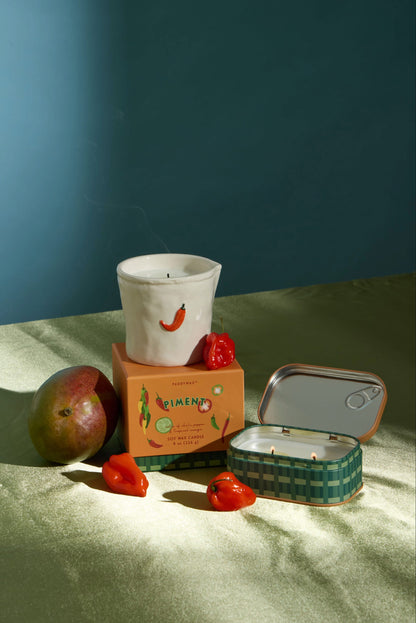 A white ceramic fruit and spice candle, an orange box, mango, chili peppers, and the Piment - Bistro 4.5 oz Tin Candle are arranged on a green surface under soft lighting against a blue-green background. Subtle hints of the Piment fragrance linger in the air, evoking warmth and vibrancy throughout the scene.