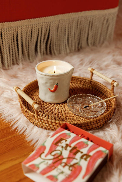 A lit candle in a white cup with Extra Hot - Square Boxed Matches rests on a wicker tray beside a glass dish and safety matches. A book titled Extra sits nearby—a perfect host gift.