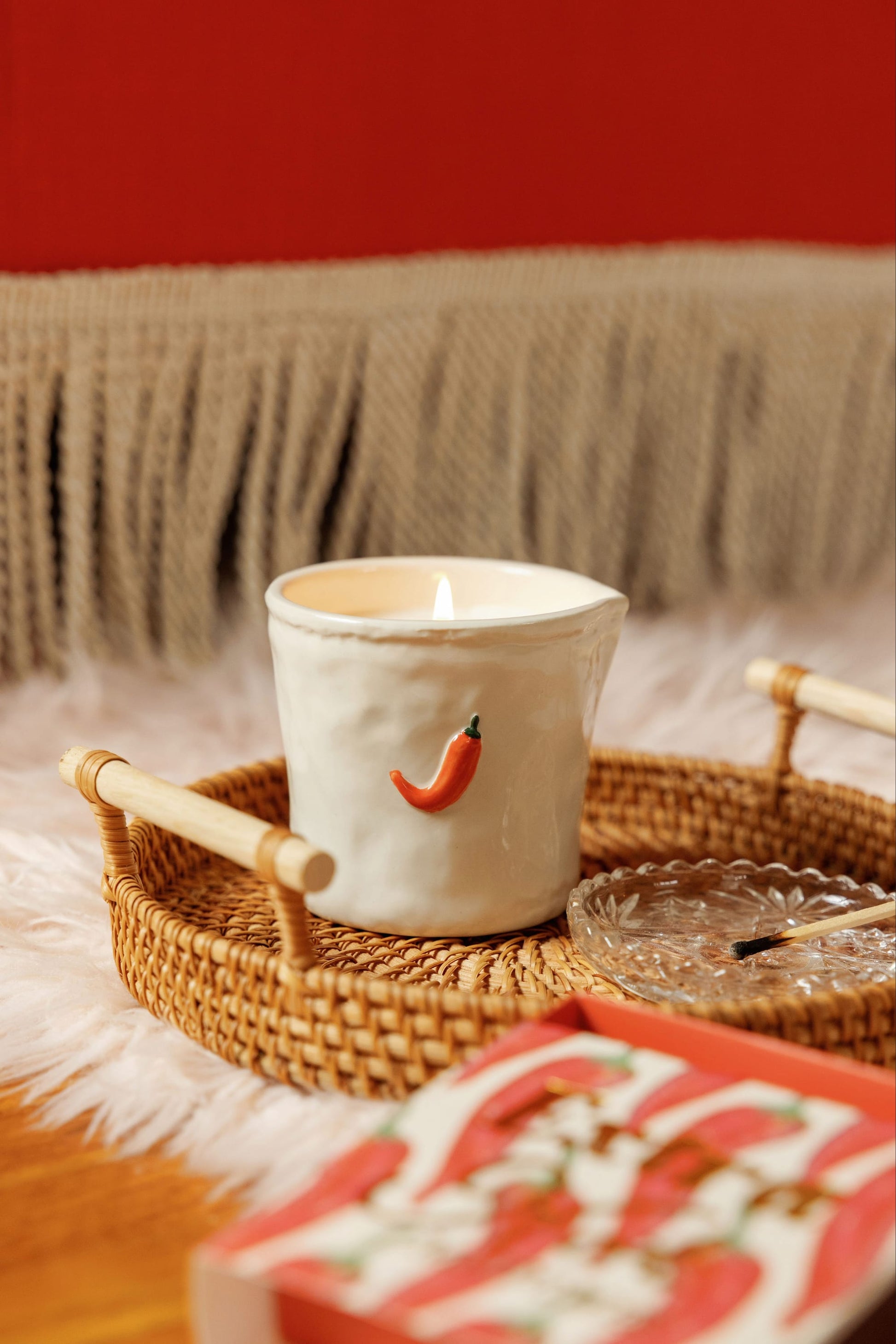The Piment - Bistro 8 oz Candle, housed in a white ceramic vessel featuring a chili pepper motif, glows brightly as it rests on a woven tray beside a glass ashtray. Its subtle fragrance fills the air, while a fringed blanket and a red wall create an inviting backdrop.