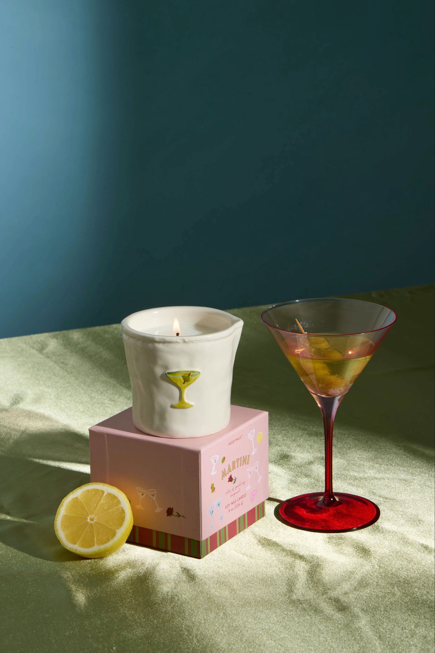 The Martini - Bistro 8 oz Candle, a lit white candle in a cup featuring a martini glass sketch, sits atop a pink box beside a red-stemmed martini glass filled with a drink and lemon twist, and a lemon slice, all arranged on a green surface against a blue and shadowed background.