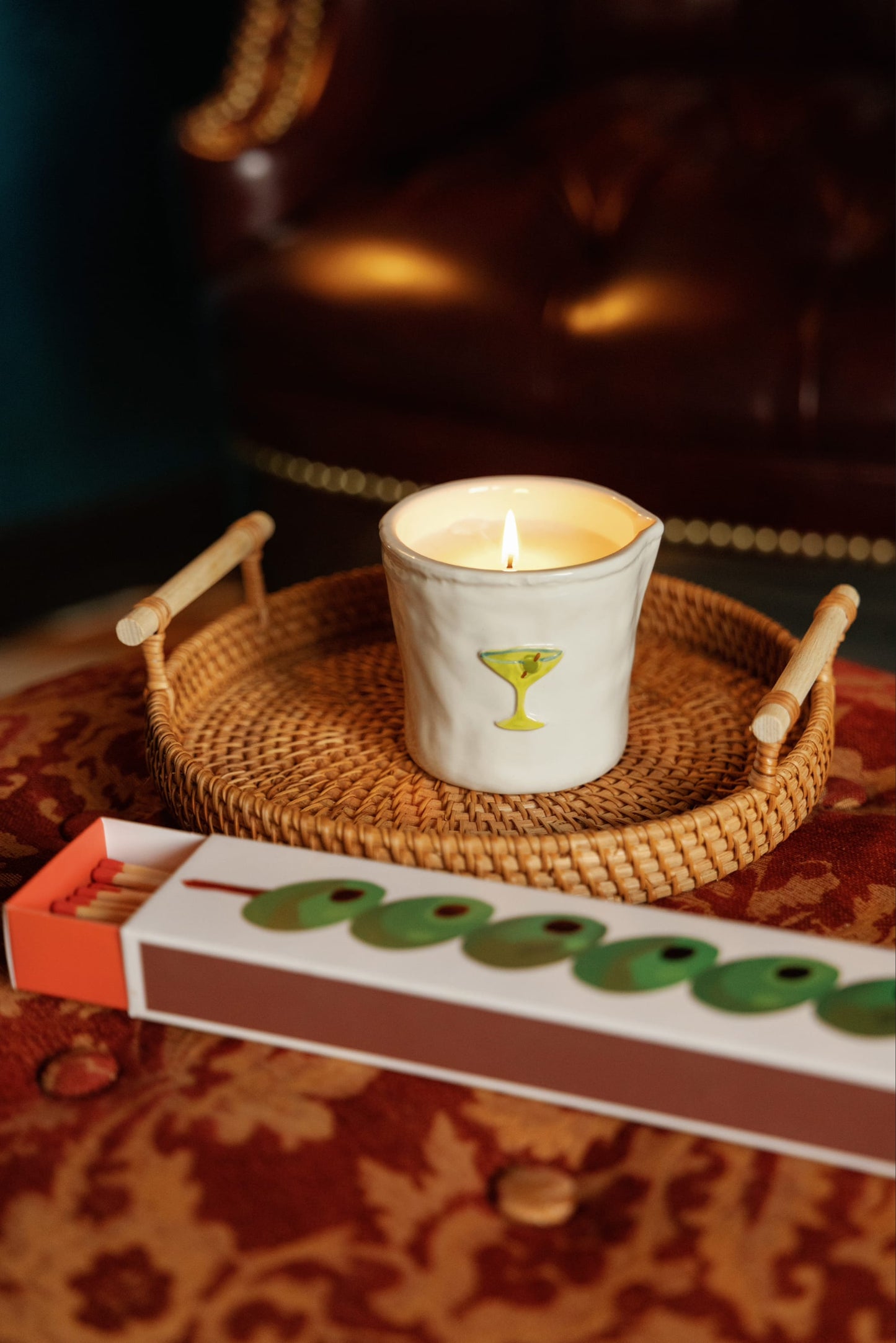 The Martini - Bistro 8 oz Candle, in a white holder featuring a martini glass design, sits on a woven tray beside a box of green-tipped matches atop a patterned surface, with a brown leather chair in the background.