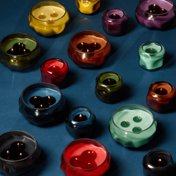 Colorful glass votive candle holders with lit candles are arranged on a dark blue surface. The holders vary in size, shape, and color, each containing one or more candle flames.