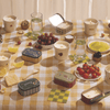 A checkered tablecloth with candles in tins, bowls of fruit, glasses of water, lemon slices, crackers, tomatoes, a cupcake, and small dishes arranged on the table.