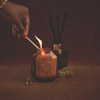 A hand lights a brown glass candle with a match; a reed diffuser and some dried herbs are in the background on a brown surface.