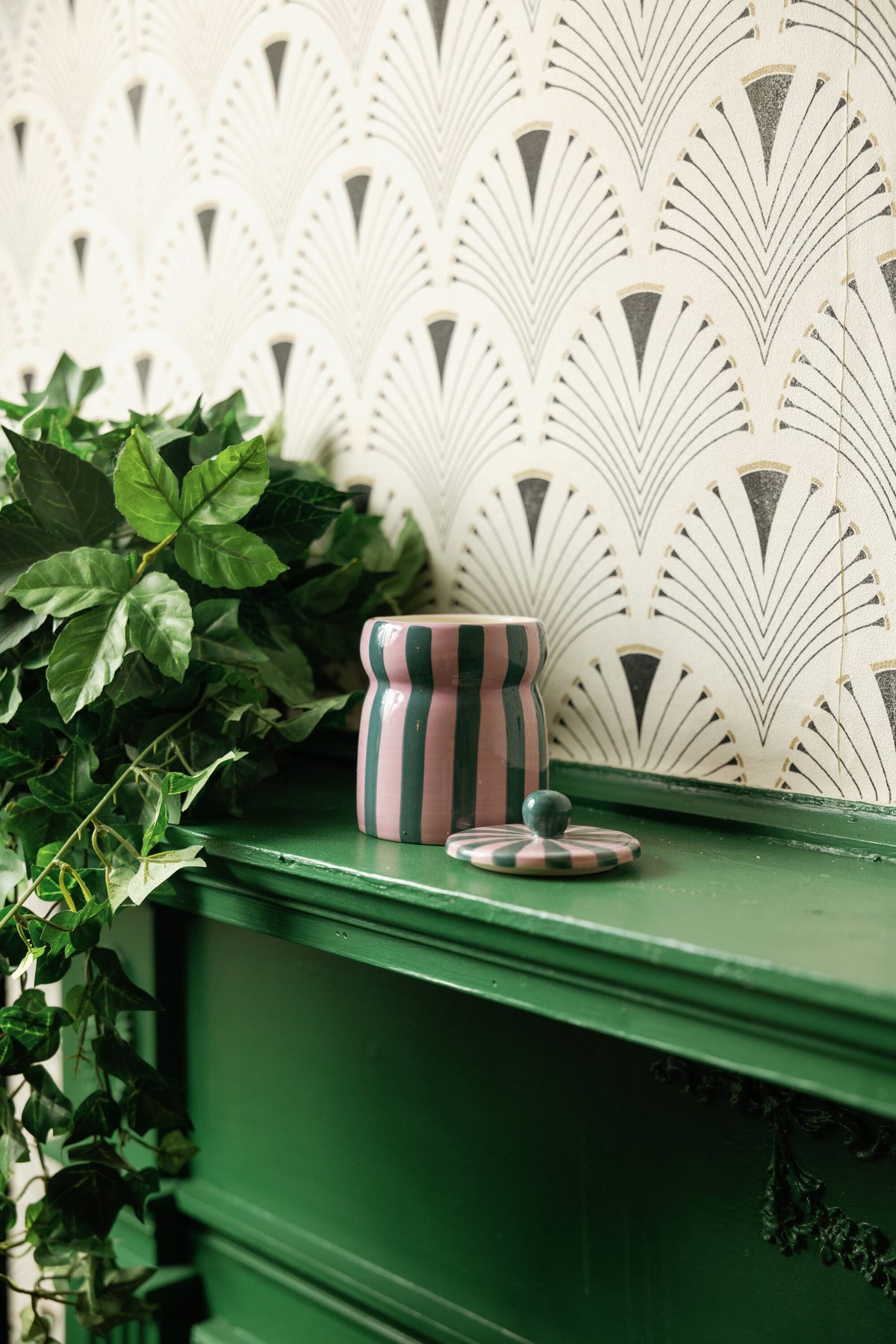 A Cinnamon & Clove - Cabana 6.5 oz Candle, featuring a pink and green striped ceramic jar, sits on a green shelf beside some leafy green plants, with art deco patterned wallpaper in the background.