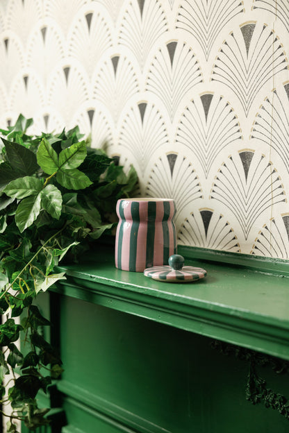 A Cinnamon & Clove - Cabana 6.5 oz Candle, featuring a pink and green striped ceramic jar, sits on a green shelf beside some leafy green plants, with art deco patterned wallpaper in the background.