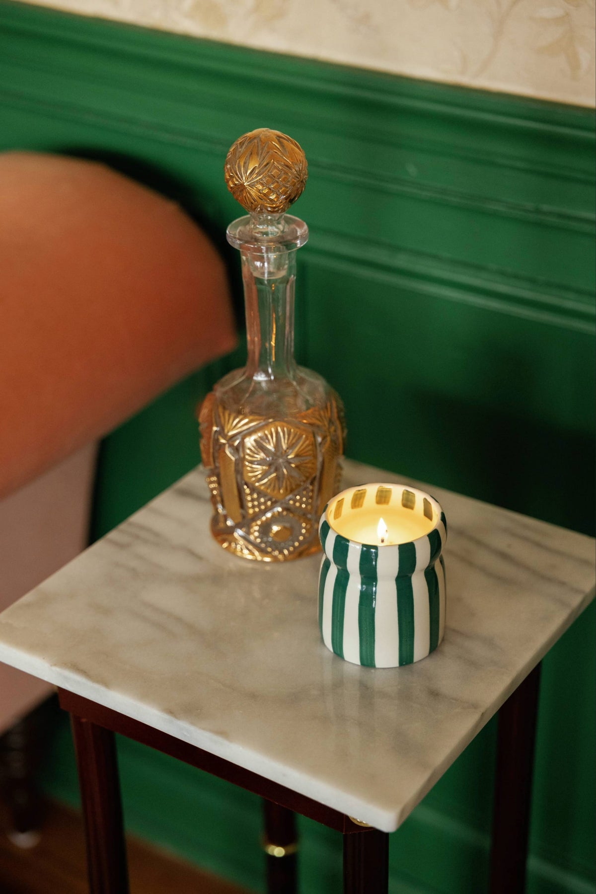 The Isla Fresca - Cabana 6.5 oz Candle, its flame casting a warm glow, sits alongside a decorative glass decanter adorned with ornate gold accents on a small marble-topped table beside a green paneled wall and an upholstered chair.