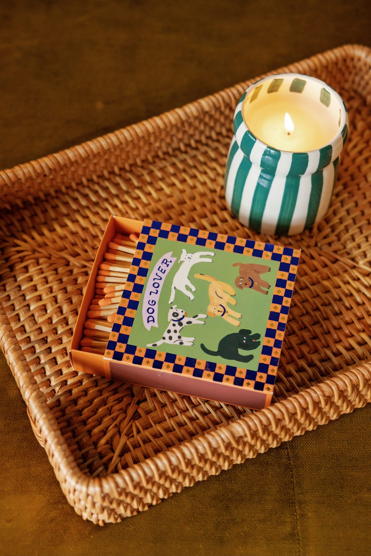 A lit Isla Fresca - Cabana 6.5 oz Candle in a green and white striped holder is placed next to a box of matches decorated with cartoon dogs and the words “Dog Lover,” all arranged on a woven tray.