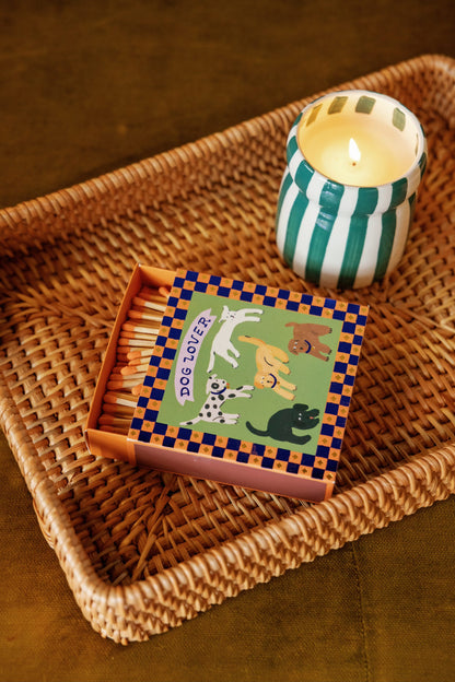 A lit Isla Fresca - Cabana 6.5 oz Candle in a green and white striped holder is placed next to a box of matches decorated with cartoon dogs and the words “Dog Lover,” all arranged on a woven tray.