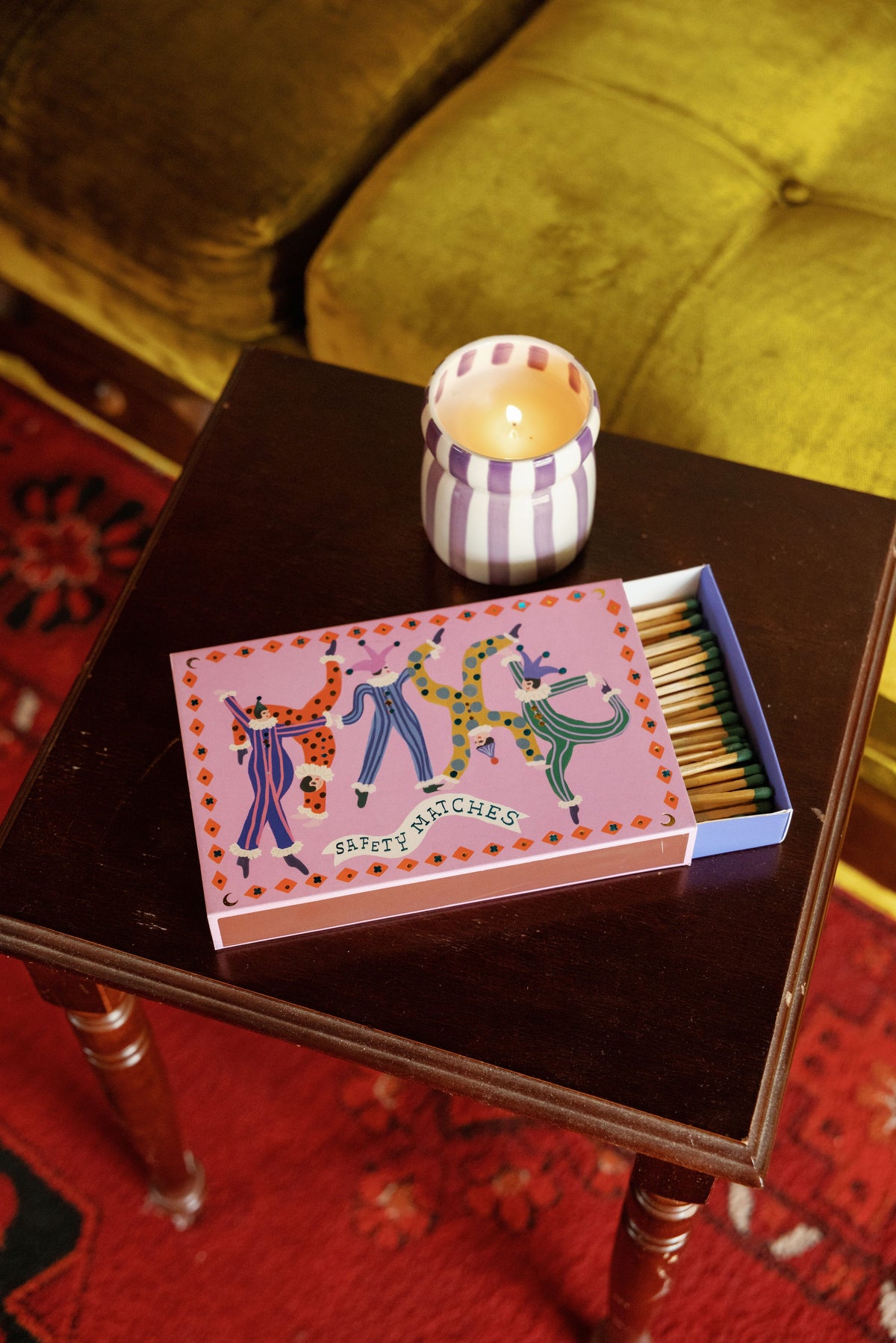 A small dark wooden table holds the Waikiki Waters - Cabana 6.5 oz Candle in a ceramic vessel alongside an open box of safety matches decorated with colorful animal designs, filling the air with its signature coastal scent. In the background, a green velvet couch and red patterned rug can be seen.